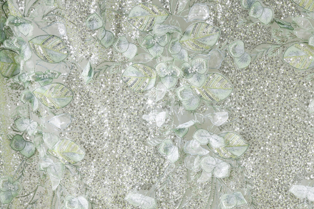 The image is a close-up of a fabric with a floral pattern, featuring a mix of green and white elements. The fabric appears to be made of sequins or glitter, adding a sparkly texture to the overall design.