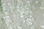 The image is a close-up of a fabric with a floral pattern, featuring a mix of green and white elements. The fabric appears to be made of sequins or glitter, adding a sparkly texture to the overall design.