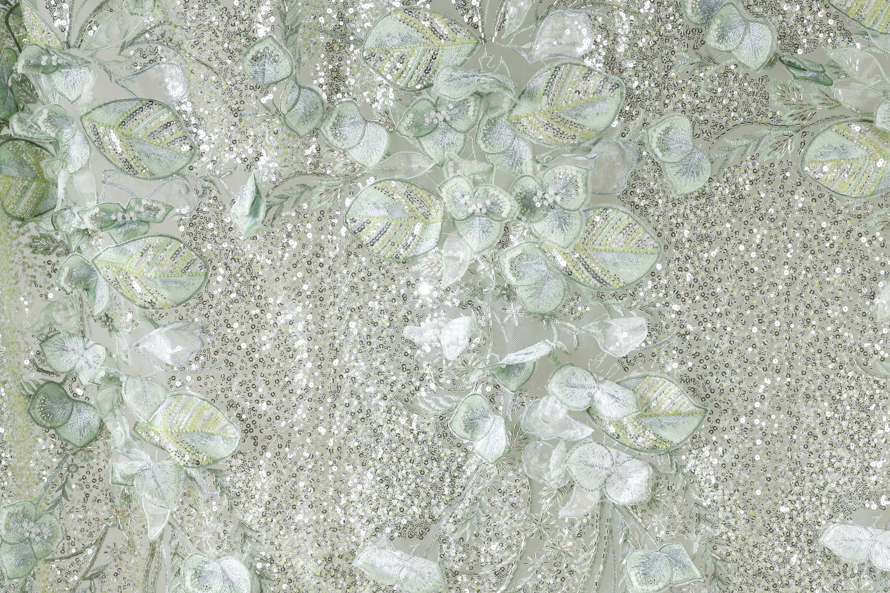 The image is a close-up of a fabric with a floral pattern, featuring a mix of green and white elements. The fabric appears to be made of sequins or glitter, adding a sparkly texture to the overall design.