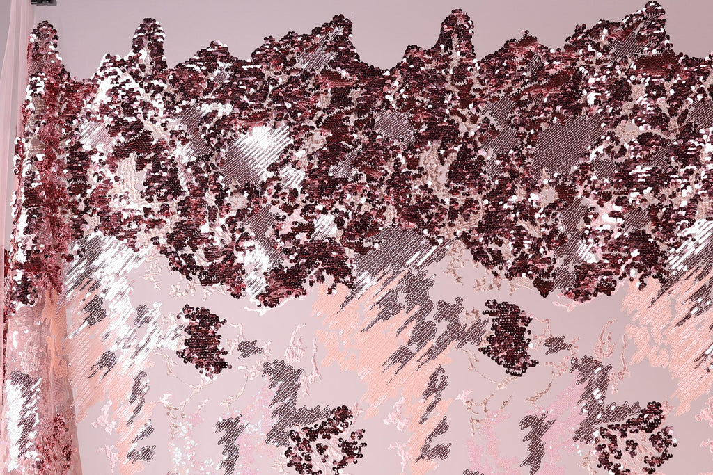 The image is a close-up of a fabric with a pink and red sequin pattern. The fabric appears to be a sheer material, with a textured and layered appearance. The pattern consists of various shades of pink and red sequins, creating a visually striking and intricate design.