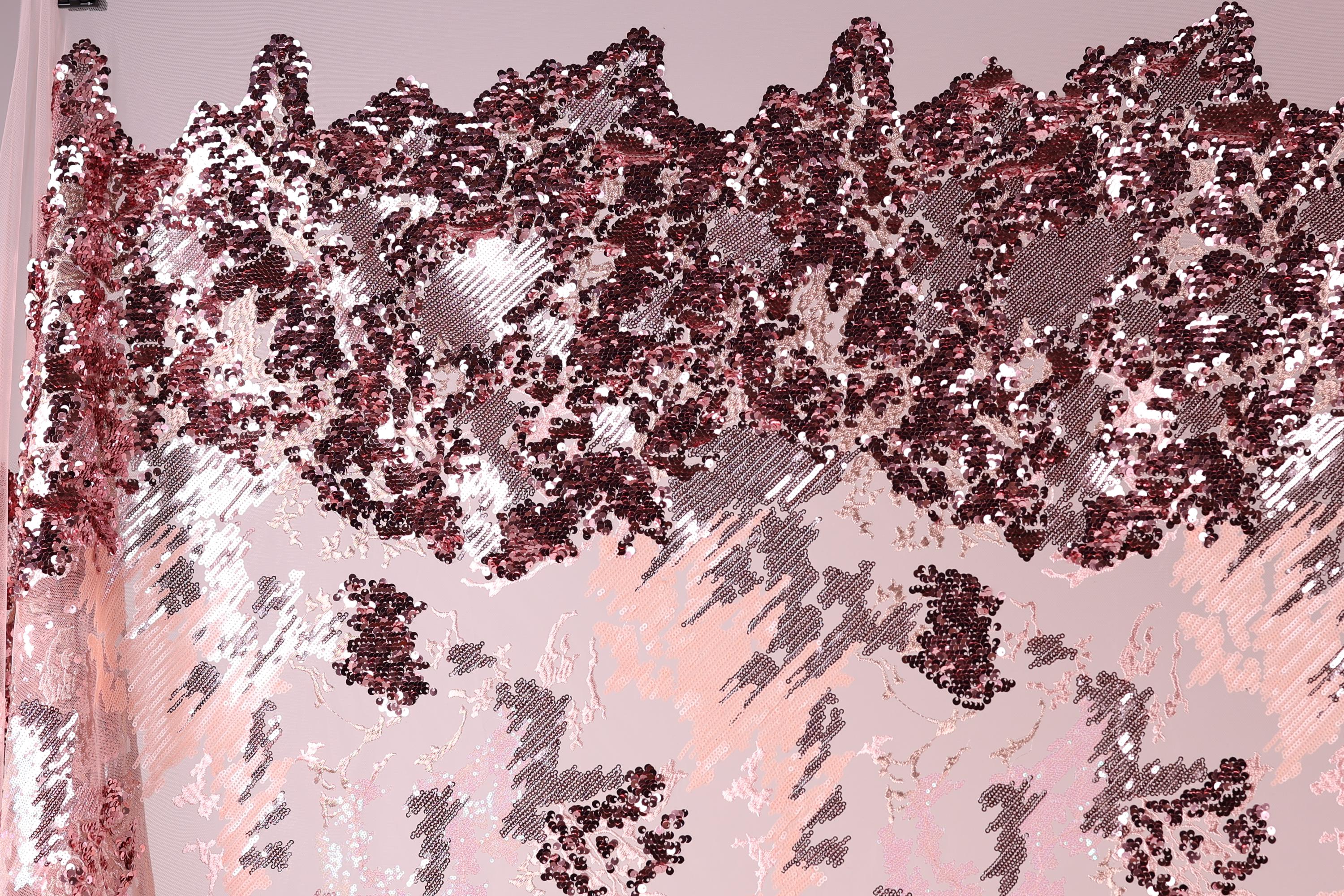 The image is a close-up of a fabric with a pink and red sequin pattern. The fabric appears to be a sheer material, with a textured and layered appearance. The pattern consists of various shades of pink and red sequins, creating a visually striking and intricate design.