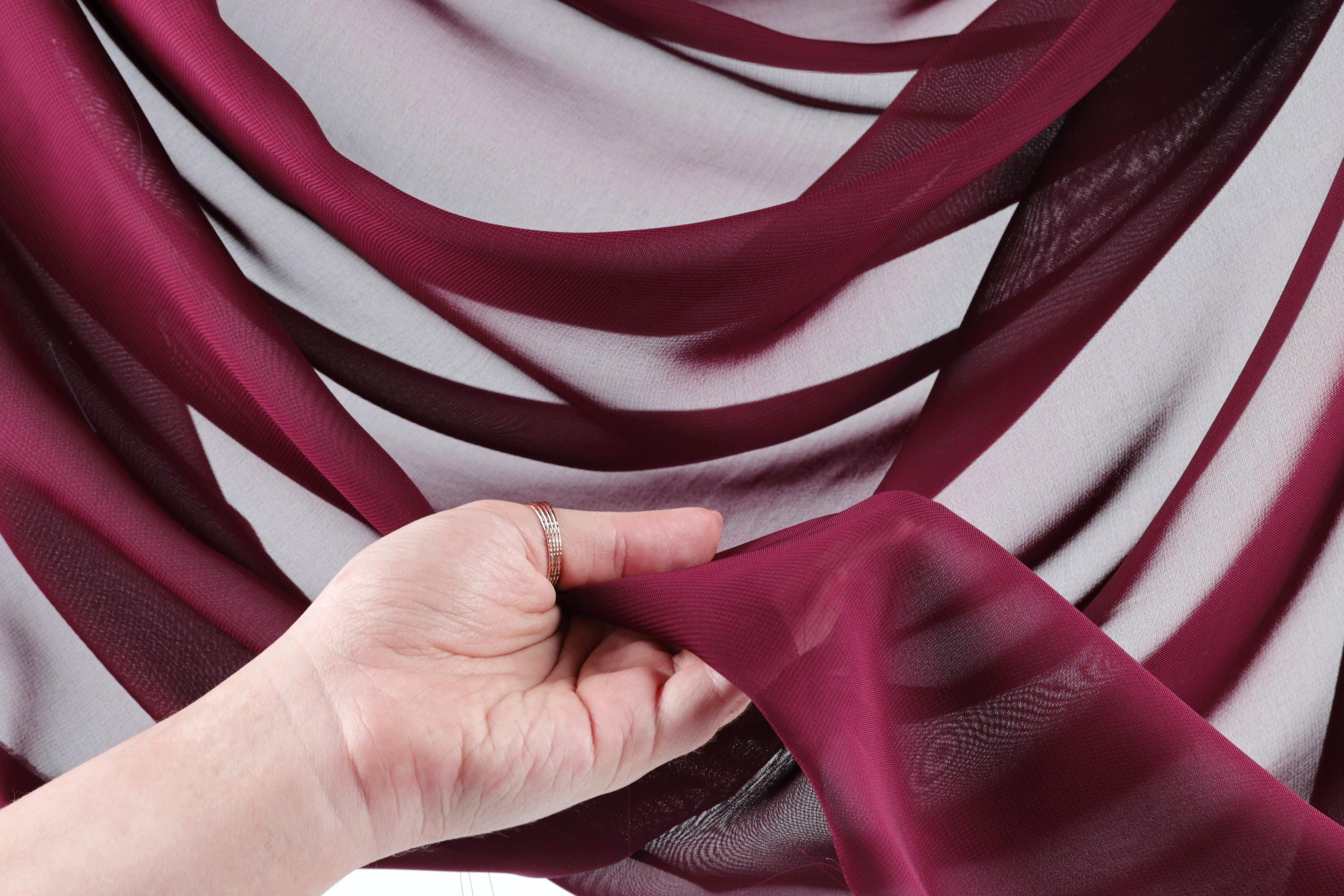 A hand is holding a piece of maroon fabric with a ring on it, against a background of white and burgundy fabric.