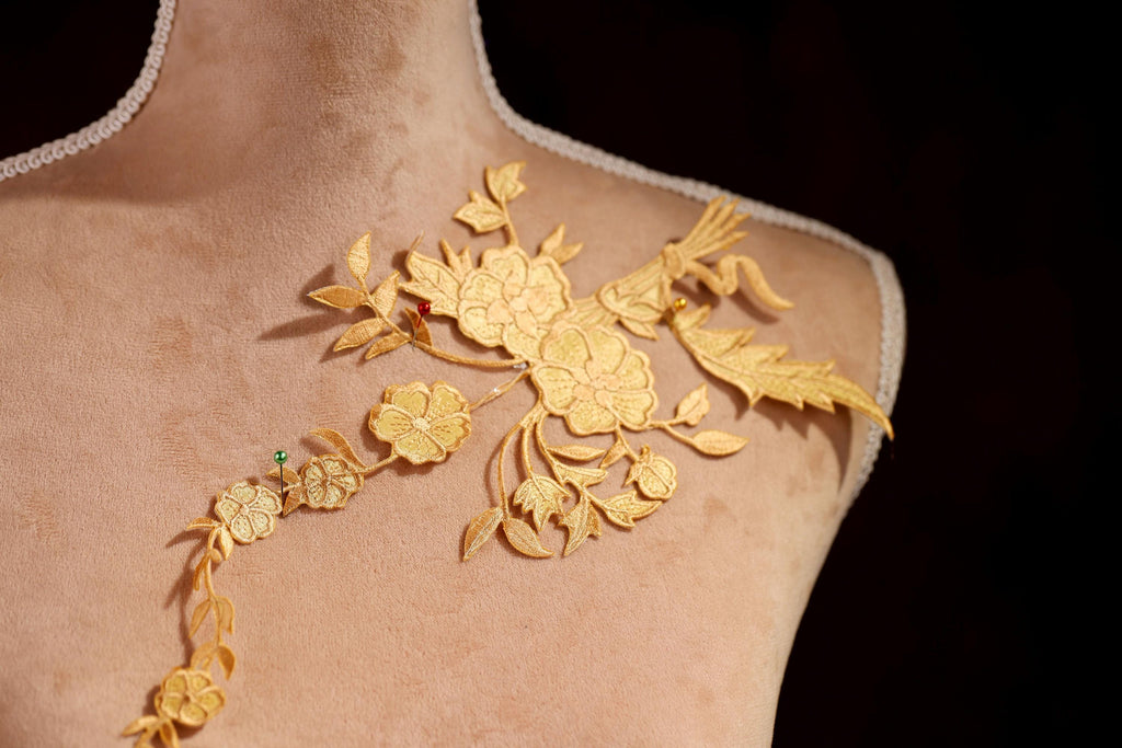 A close-up view of a gold embroidered design on a mannequin's shoulder, featuring intricate floral patterns and leaves.