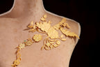 A close-up view of a gold embroidered design on a mannequin's shoulder, featuring intricate floral patterns and leaves.