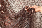A hand is holding a piece of sequin fabric, which appears to be a dark brown color with a pattern of small dots and lines. The fabric is being held up against a background that is a light gray color with a repeating pattern of lines and dots.