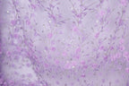 a close-up view of a fabric with a delicate floral pattern in shades of purple and white. The fabric appears to be made of a sheer material, with numerous small flowers and leaves intricately woven into the design. The overall color scheme of the fabric is predominantly purple, with accents of white and silver adding depth and contrast to the pattern.