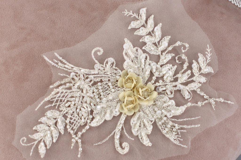 a delicate and intricate piece of lace or beading, adorned with a floral design. The design includes a central flower surrounded by smaller flowers and leaves, creating a visually appealing and elegant piece of textile art.