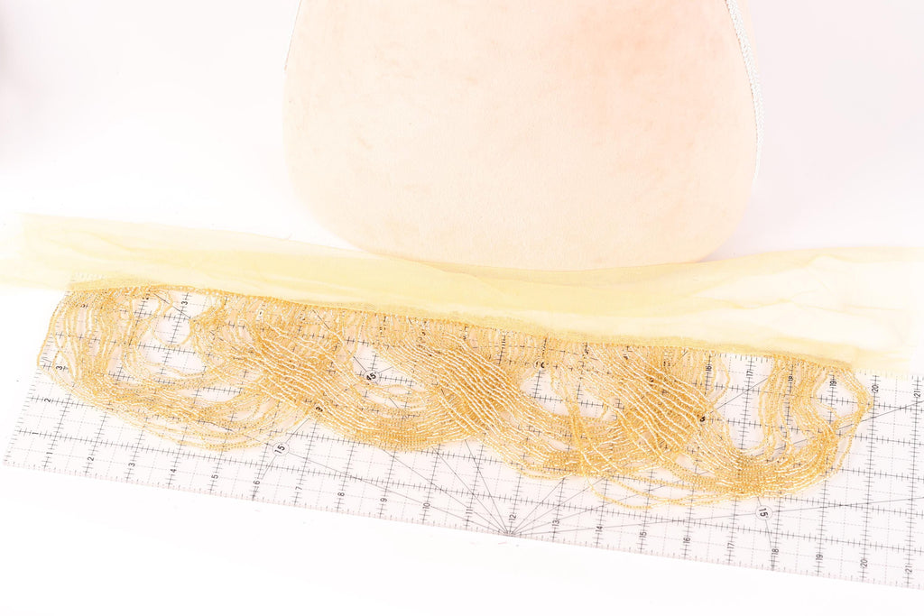 a piece of fabric with a yellow fringe, placed on a white surface. The fabric appears to be made of a material that resembles lace or a similar delicate fabric. There is a ruler or measuring mat in the foreground, which is likely used for measuring or cutting the fabric.