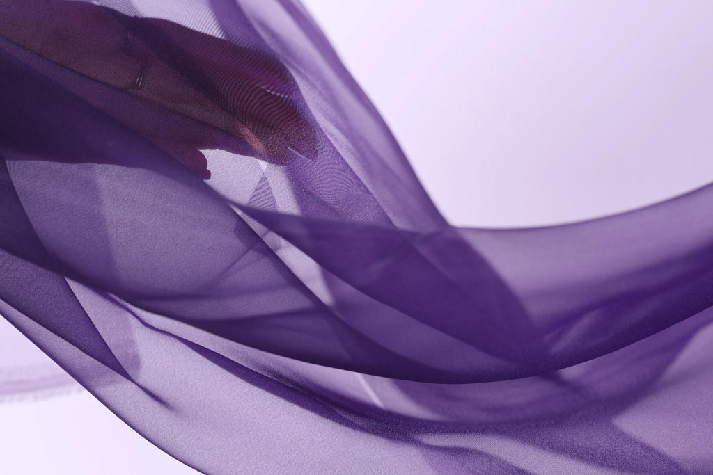 The image is a close-up of a hand holding a purple fabric, with the fabric appearing to be flowing and draped. The background is a light purple color, creating a soft and dreamy atmosphere.
