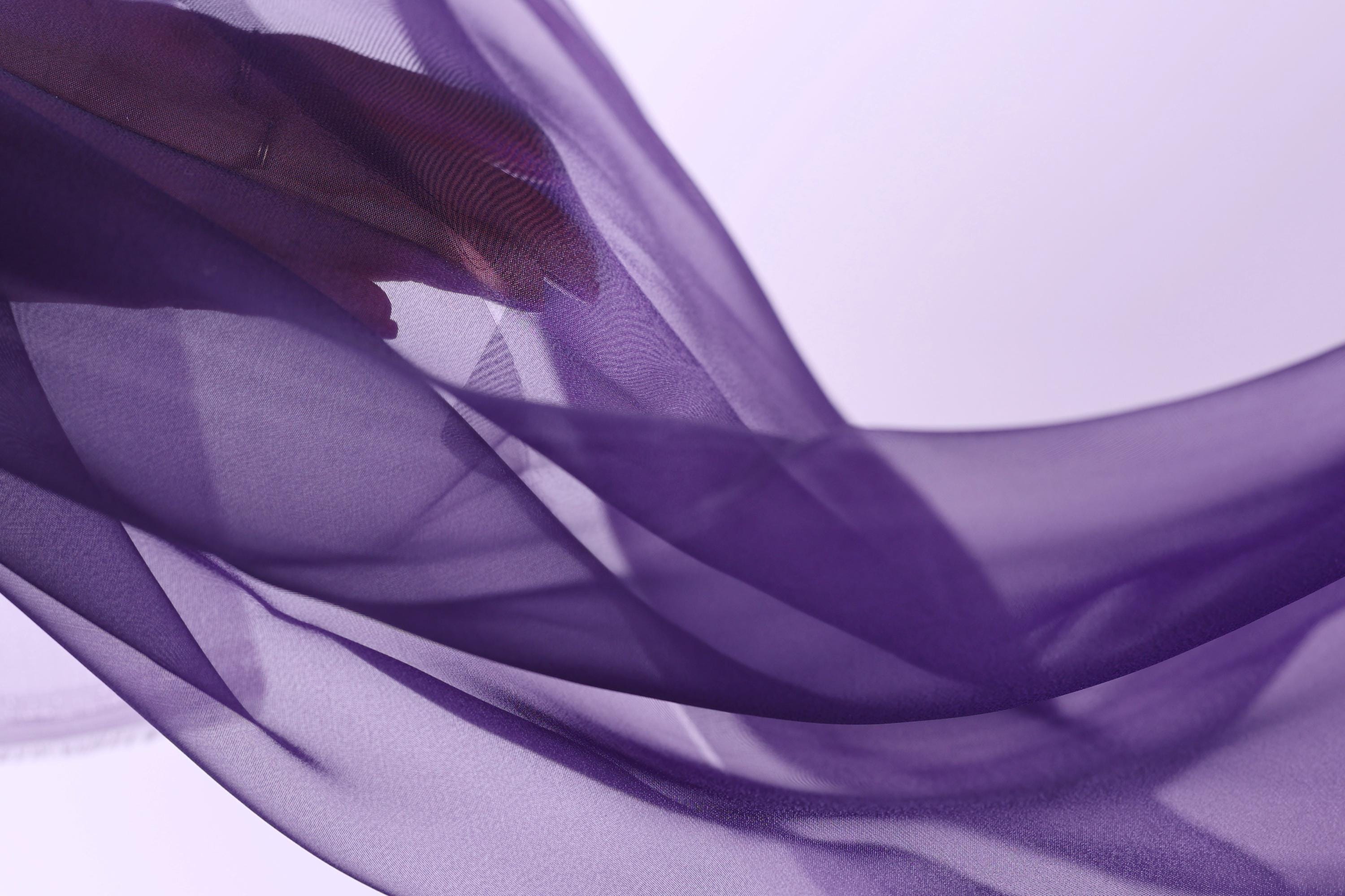The image is a close-up of a hand holding a purple fabric, with the fabric appearing to be flowing and draped. The background is a light purple color, creating a soft and dreamy atmosphere.