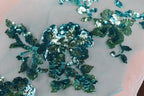a close-up view of a delicate, intricate embroidered design on a light-colored fabric. The design is composed of various shades of blue and green sequins, creating a visually striking and detailed pattern. The fabric appears to be a light pink or white material, which contrasts with the vibrant colors of the sequins and enhances their visual impact. The image does not contain any text or other discernible objects, and the focus is solely on the detailed embroidered design.