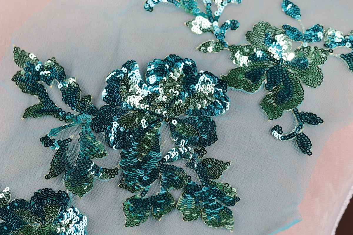 a close-up view of a delicate, intricate embroidered design on a light-colored fabric. The design is composed of various shades of blue and green sequins, creating a visually striking and detailed pattern. The fabric appears to be a light pink or white material, which contrasts with the vibrant colors of the sequins and enhances their visual impact. The image does not contain any text or other discernible objects, and the focus is solely on the detailed embroidered design.