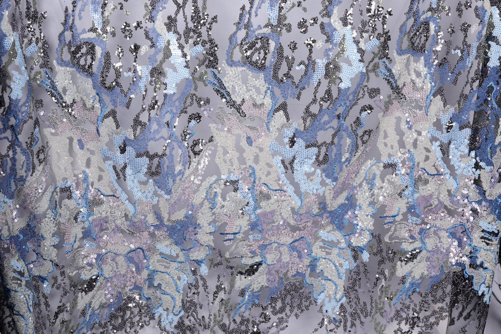a close-up view of a fabric with a complex, abstract pattern of sequins and threads in shades of blue, gray, and white. The pattern appears to be a blend of various textures and colors, creating a visually striking and intricate design.