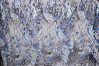 a close-up view of a fabric with a complex, abstract pattern of sequins and threads in shades of blue, gray, and white. The pattern appears to be a blend of various textures and colors, creating a visually striking and intricate design.
