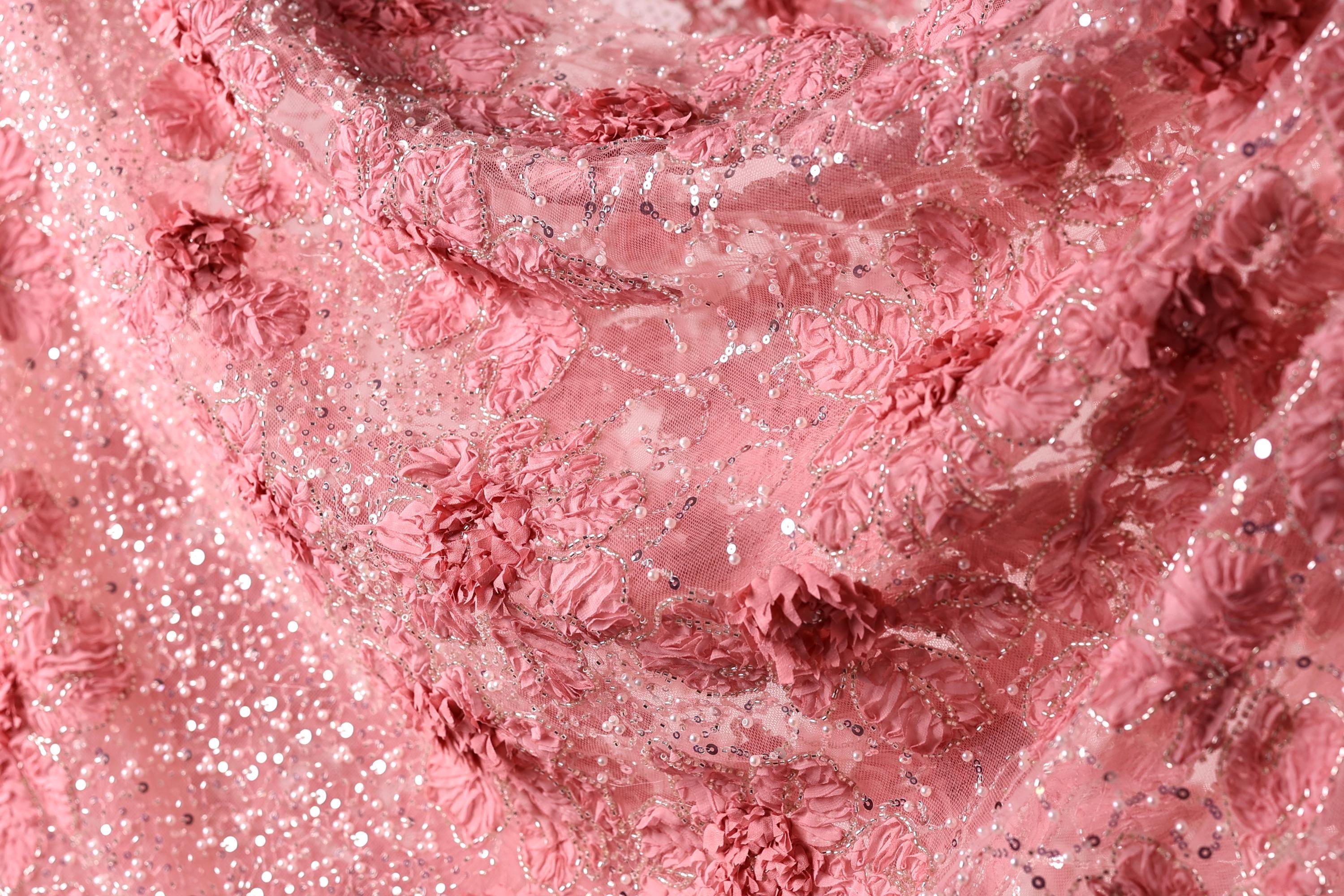 The image is a close-up of a fabric with a pink floral pattern, featuring numerous small pink flowers and sequins or beads.