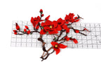 a close-up view of a red flower with a yellow center, surrounded by a branch with red flowers and green leaves. The flower is placed on a ruler, which is placed on a white surface.