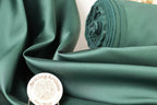 a close-up of a green satin fabric, with a rolled-up spool of the same material visible in the background. There is also a small white circular object with text on it placed in the foreground.