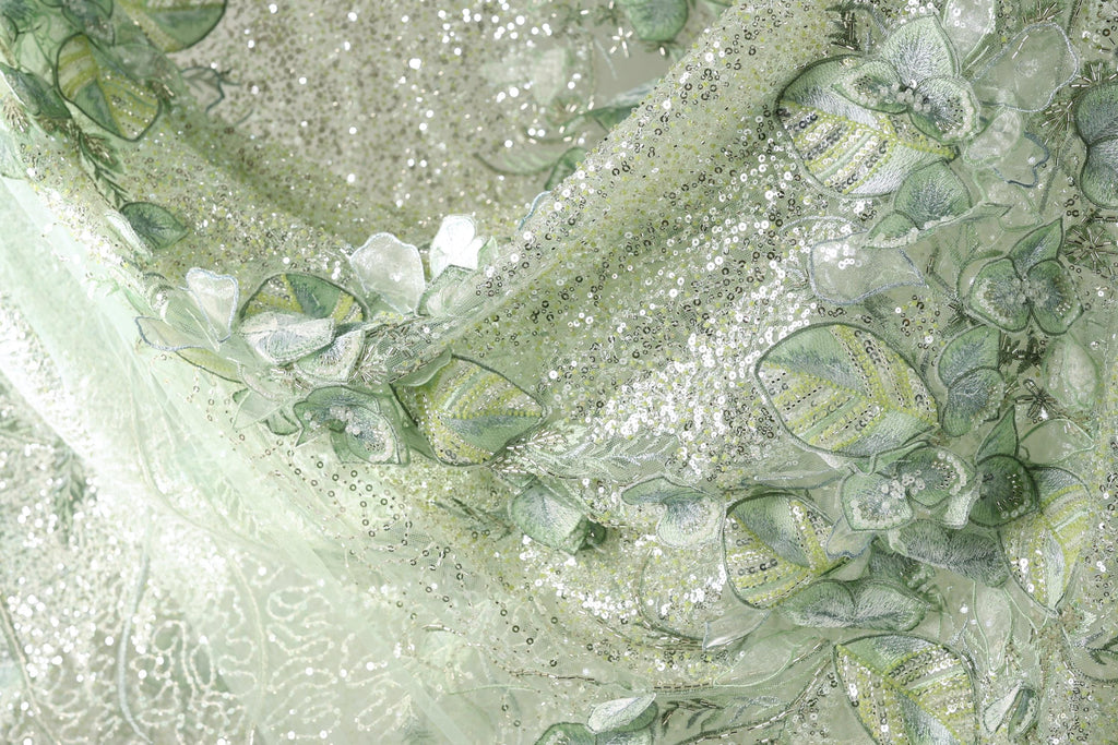 The image is a close-up of a fabric with a floral pattern, featuring green and white colors and small sequins or glitter accents.