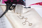 a close-up view of a piece of jewelry, including a necklace with a large, sparkling crystal pendant, placed on a white fabric surface. There are also some scissors and a book visible in the scene.