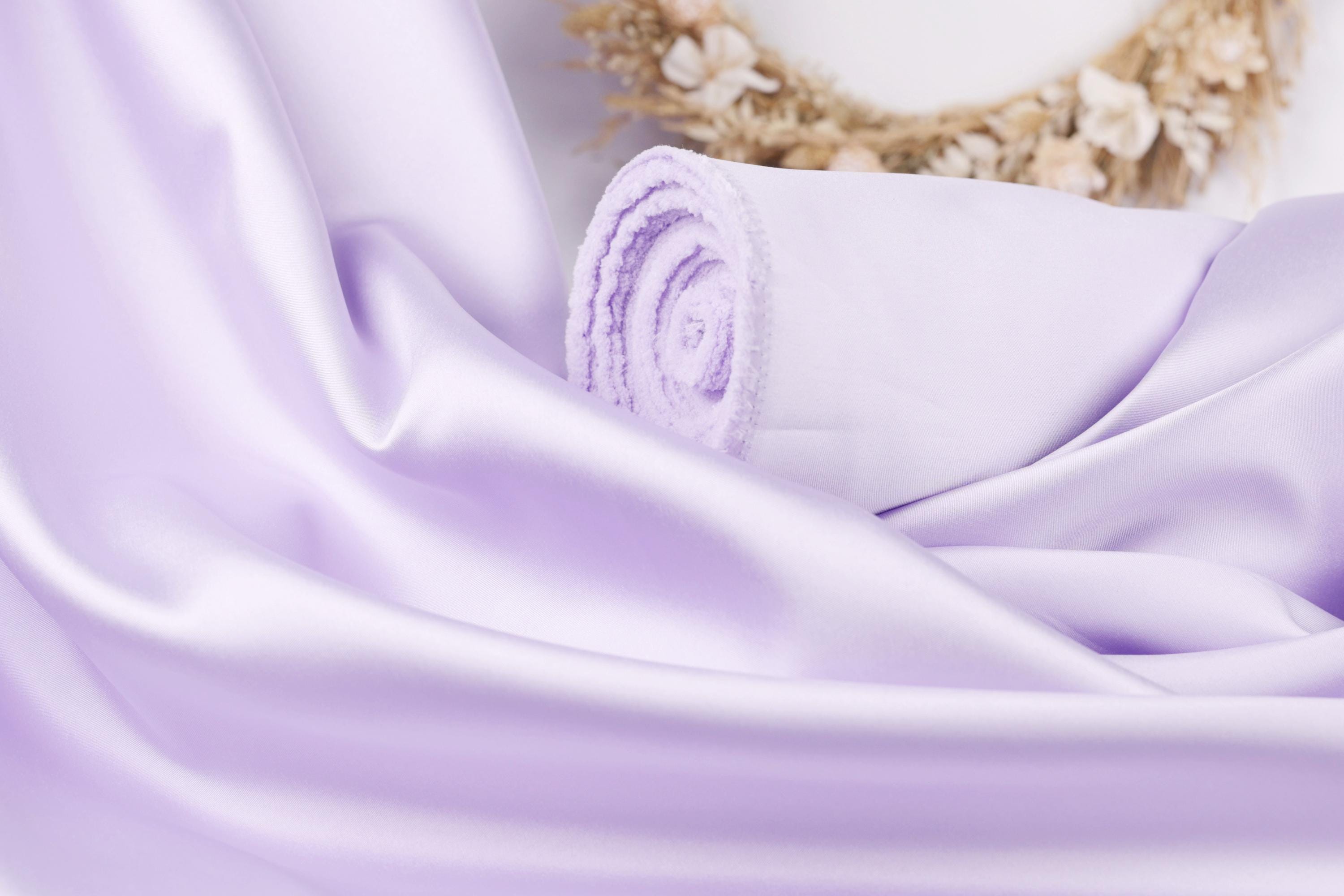 A close-up view of a soft, flowing purple fabric, possibly a satin or satin-like material, with a curled edge visible. In the background, there is a wreath made of dried flowers, adding a natural element to the composition.