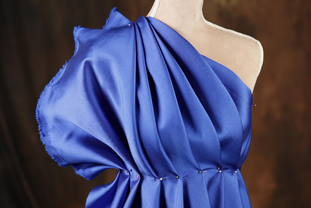 A close-up view of a blue one-shoulder dress with a draped, flowing fabric and a ruched neckline.
