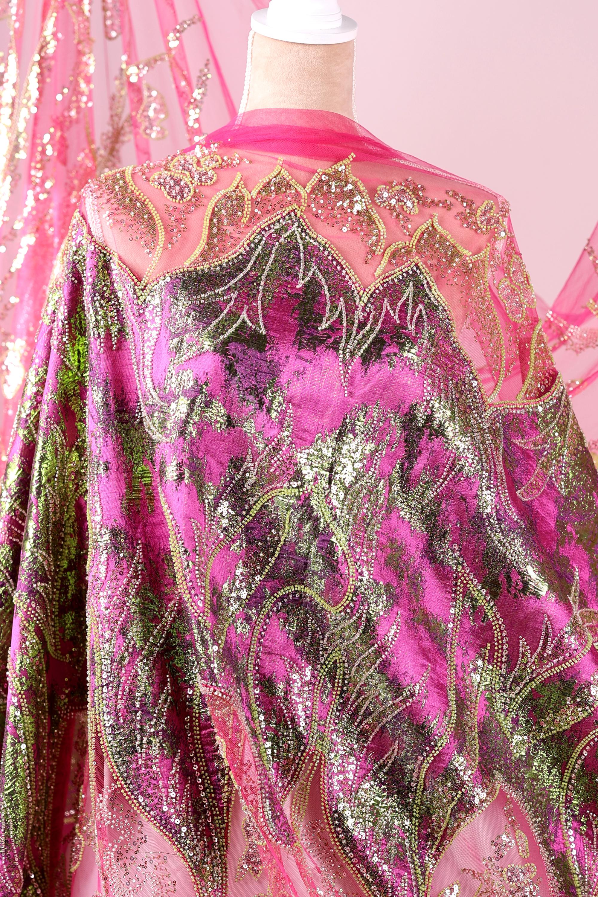 a vibrant, intricately designed fabric with a mix of pink, green, and gold colors. The fabric appears to be a sheer material, with a floral pattern and sequin accents. The design is detailed and eye-catching, showcasing the craftsmanship and artistry involved in creating this piece of clothing or textile.