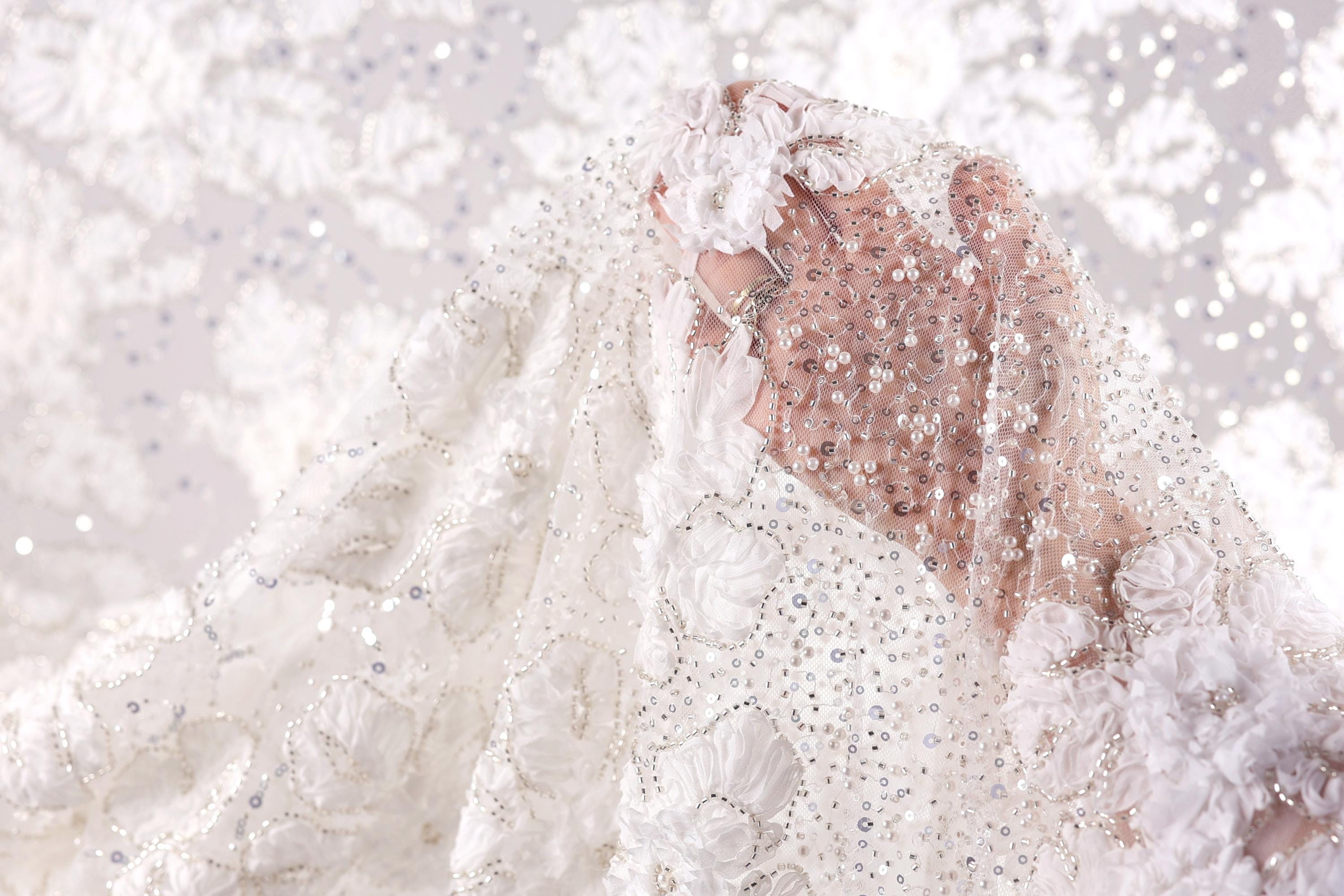 A close-up view of a white wedding dress with intricate beading and lace details, partially obscured by a veil.