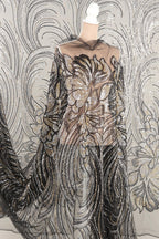 a mannequin wearing a long, flowing dress with a sheer, sequined bodice and a sheer, black and gold patterned skirt. The dress has a dramatic, voluminous silhouette and appears to be made of a sheer, black and gold fabric.