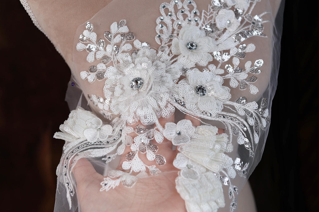 A close-up view of a delicate, intricately embroidered white lace piece, featuring a floral design with small, sparkling embellishments.