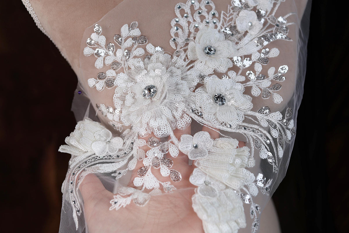 A close-up view of a delicate, intricately embroidered white lace piece, featuring a floral design with small, sparkling embellishments.