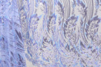a close-up view of a fabric with a repeating pattern of purple and white sequin designs. The pattern appears to be a floral or leaf-like design, with the sequins adding a touch of sparkle and texture to the fabric. The fabric is predominantly white, with the purple and white sequins creating a visually striking contrast. The image does not contain any text or other discernible objects, and the focus is solely on the intricate and detailed pattern of the fabric.