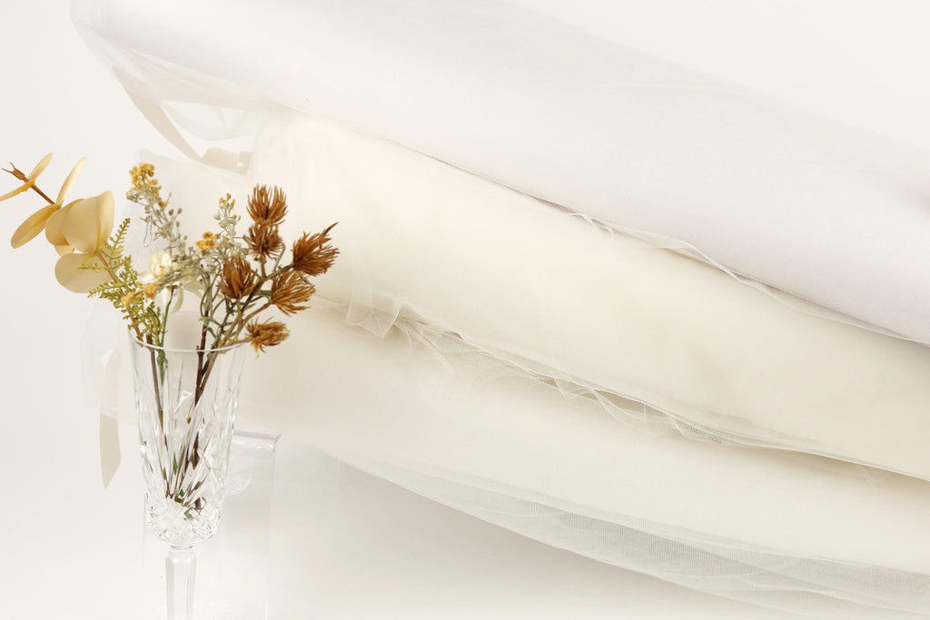 a clear glass vase filled with yellow flowers and brown dried flowers, placed on a white surface next to a white fabric or material.