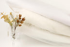 a clear glass vase filled with yellow flowers and brown dried flowers, placed on a white surface next to a white fabric or material.
