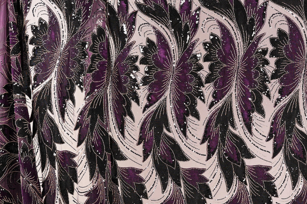 a close-up view of a fabric with a repeating floral pattern in shades of purple and black. The pattern consists of large, stylized flowers with intricate details and a mix of black and purple colors. The fabric appears to be made of a shiny, sequined material, adding a touch of sparkle and elegance to the design.