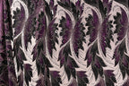 a close-up view of a fabric with a repeating floral pattern in shades of purple and black. The pattern consists of large, stylized flowers with intricate details and a mix of black and purple colors. The fabric appears to be made of a shiny, sequined material, adding a touch of sparkle and elegance to the design.