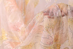 a close-up view of a delicate, sheer fabric with a floral pattern. The fabric appears to be made of a light-colored material, possibly cotton or chiffon, with a shimmering, sequined design. The fabric is being held by someone's hand, and the background is blurred, drawing attention to the fabric and its intricate details.