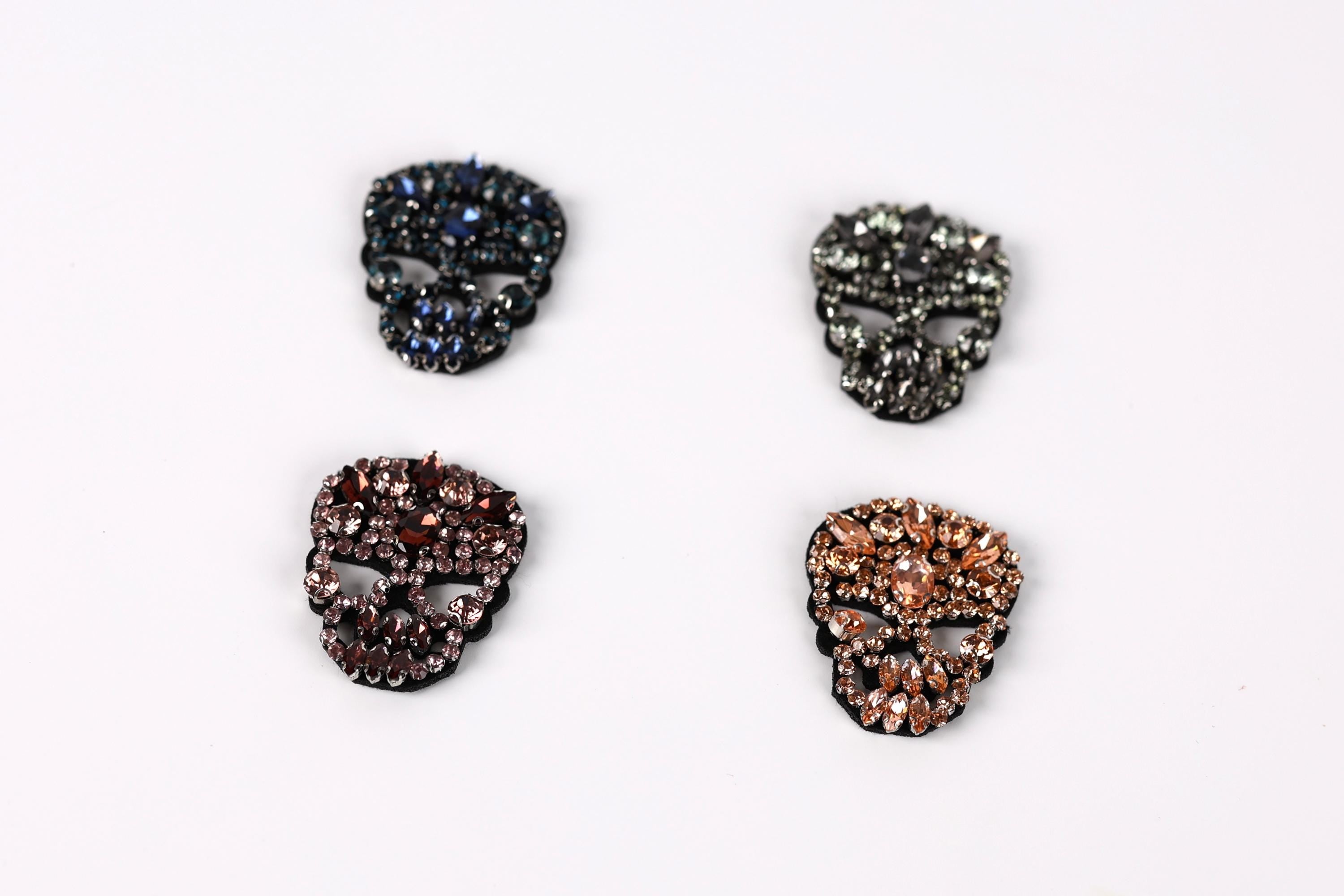 four ornate skull-shaped brooches, each with a unique color and design, arranged in a square formation against a white background.