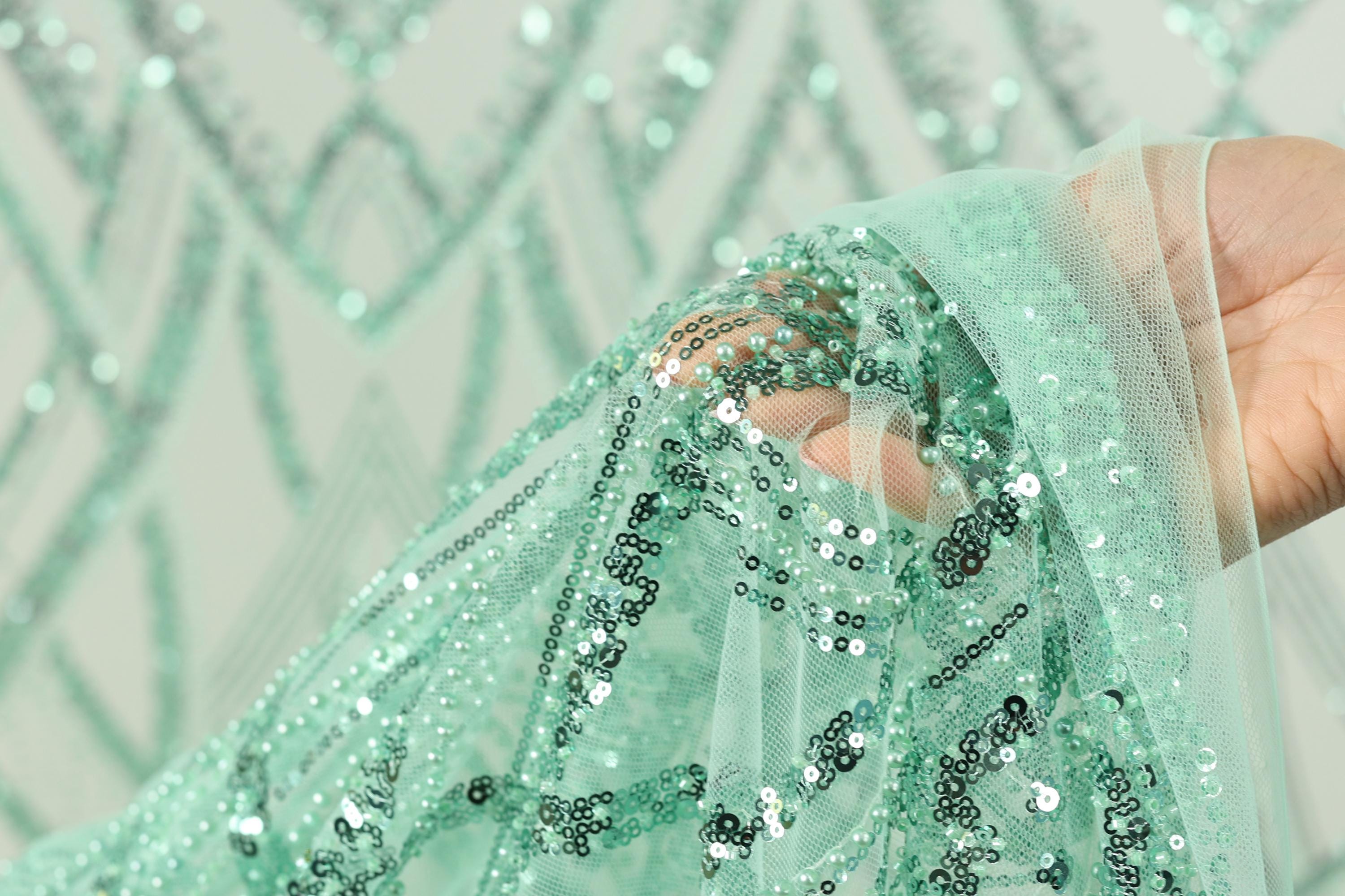 A hand is holding a piece of fabric with a green sequin pattern, which appears to be a decorative textile.