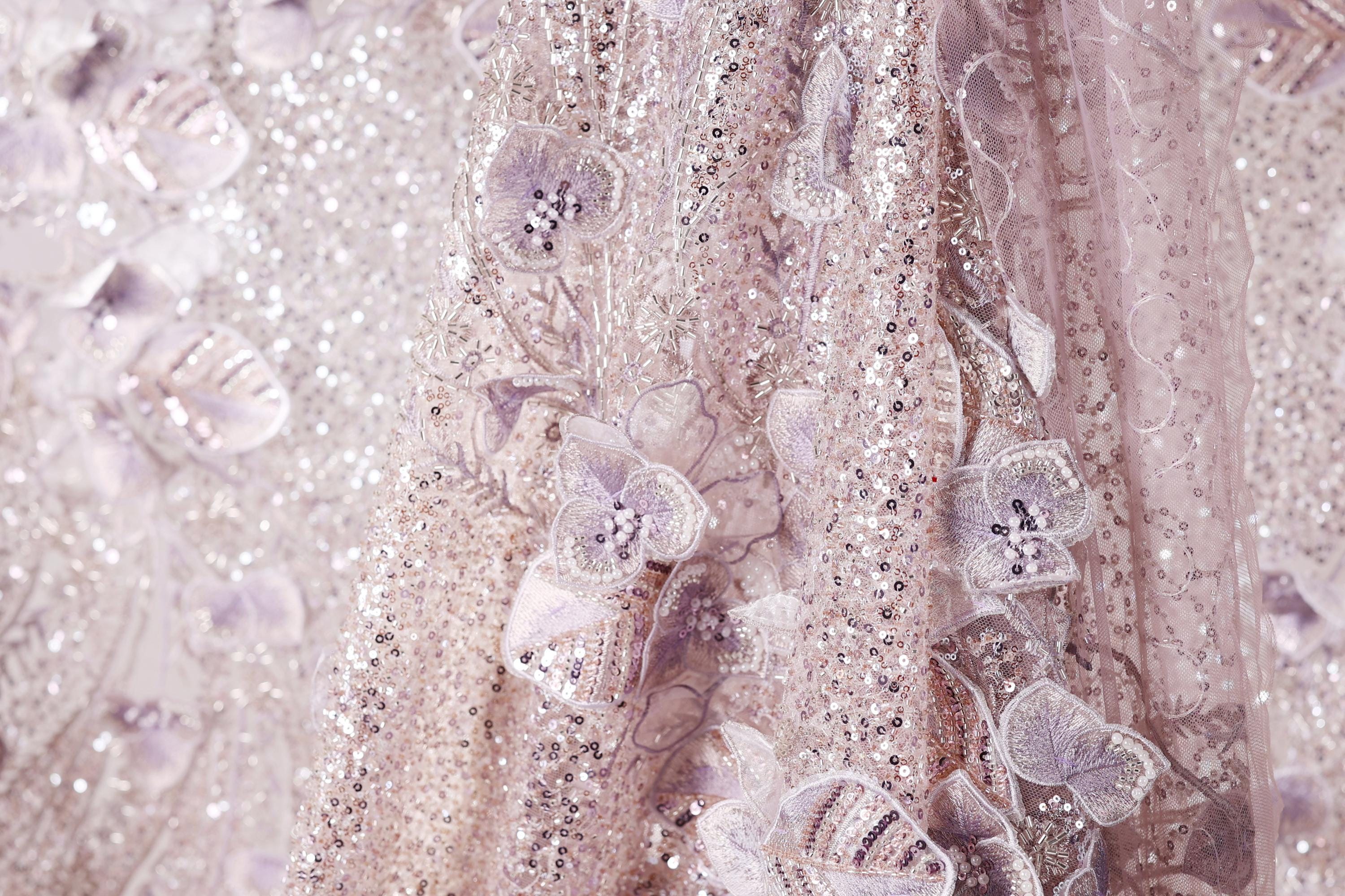 The image is a close-up of a delicate, shimmering fabric with a floral pattern. The fabric appears to be made of sequins and lace, creating a visually striking and intricate design.
