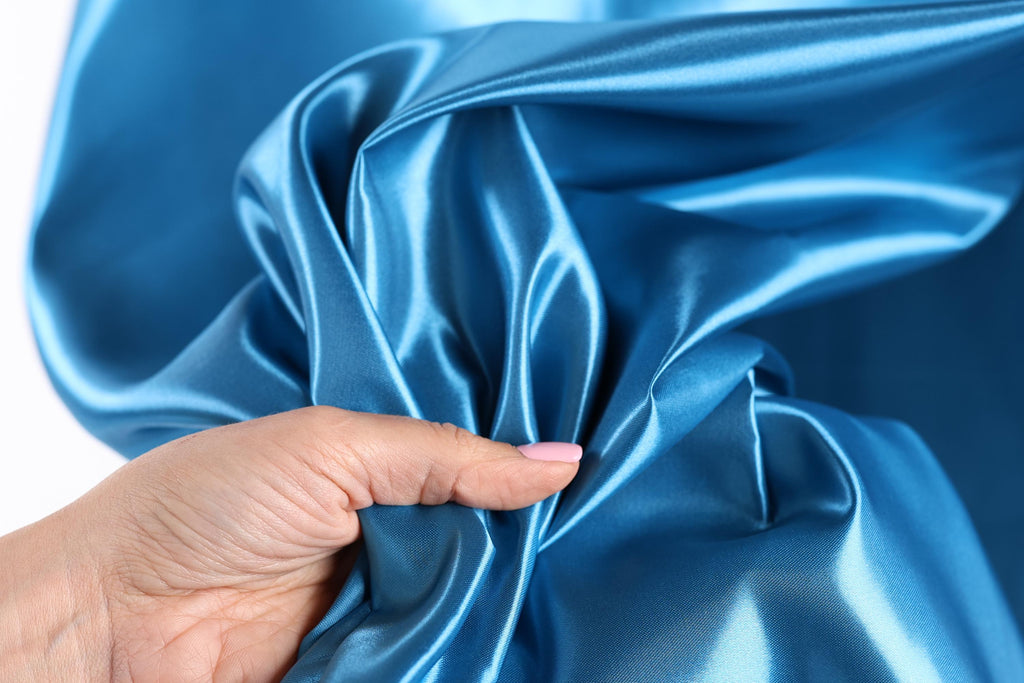 A hand is holding a piece of blue satin fabric, with the fabric's folds and creases visible.