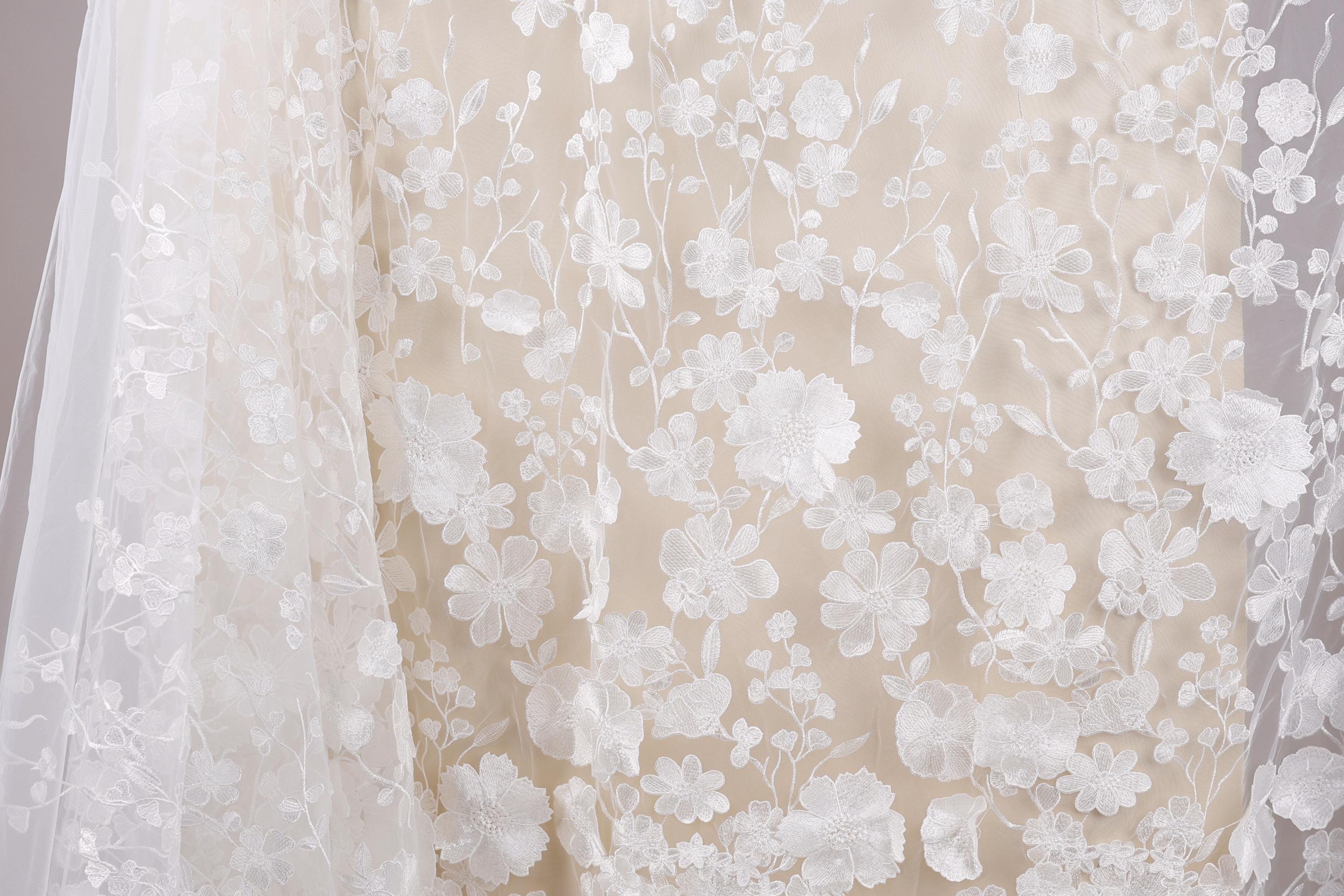 a close-up view of a delicate, white lace fabric with a floral pattern. The fabric appears to be a sheer material, with a light beige background that complements the white color of the lace. The lace is intricately designed with numerous small white flowers and leaves, creating a visually appealing and delicate appearance.