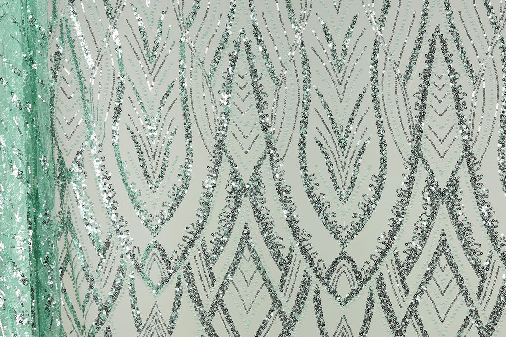 a close-up view of a fabric with a repeating pattern of silver and green sequin designs. The pattern consists of various diamond shapes and lines, creating a visually striking and intricate design. The fabric appears to be a light green color, which complements the silver and green sequins. The image does not contain any text or other discernible objects, and the focus is solely on the detailed pattern of the sequin design.