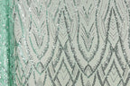 a close-up view of a fabric with a repeating pattern of silver and green sequin designs. The pattern consists of various diamond shapes and lines, creating a visually striking and intricate design. The fabric appears to be a light green color, which complements the silver and green sequins. The image does not contain any text or other discernible objects, and the focus is solely on the detailed pattern of the sequin design.