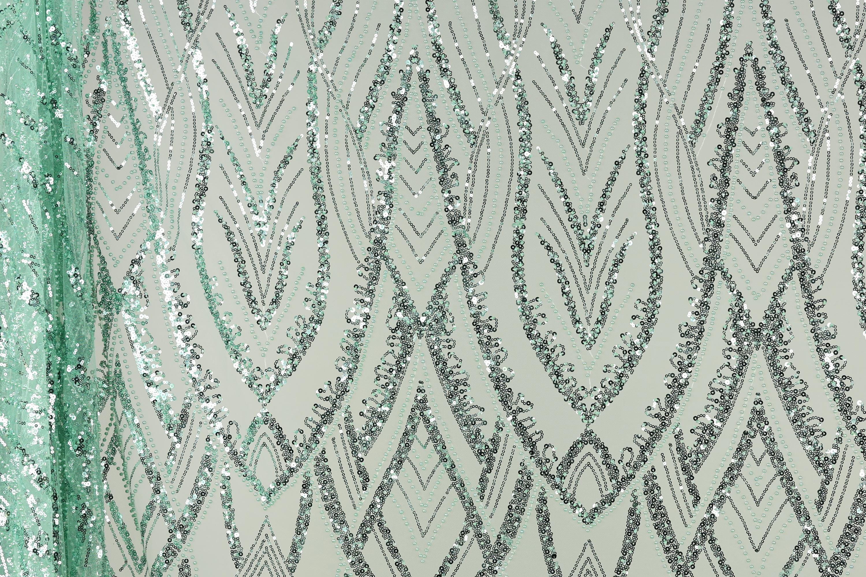a close-up view of a fabric with a repeating pattern of silver and green sequin designs. The pattern consists of various diamond shapes and lines, creating a visually striking and intricate design. The fabric appears to be a light green color, which complements the silver and green sequins. The image does not contain any text or other discernible objects, and the focus is solely on the detailed pattern of the sequin design.