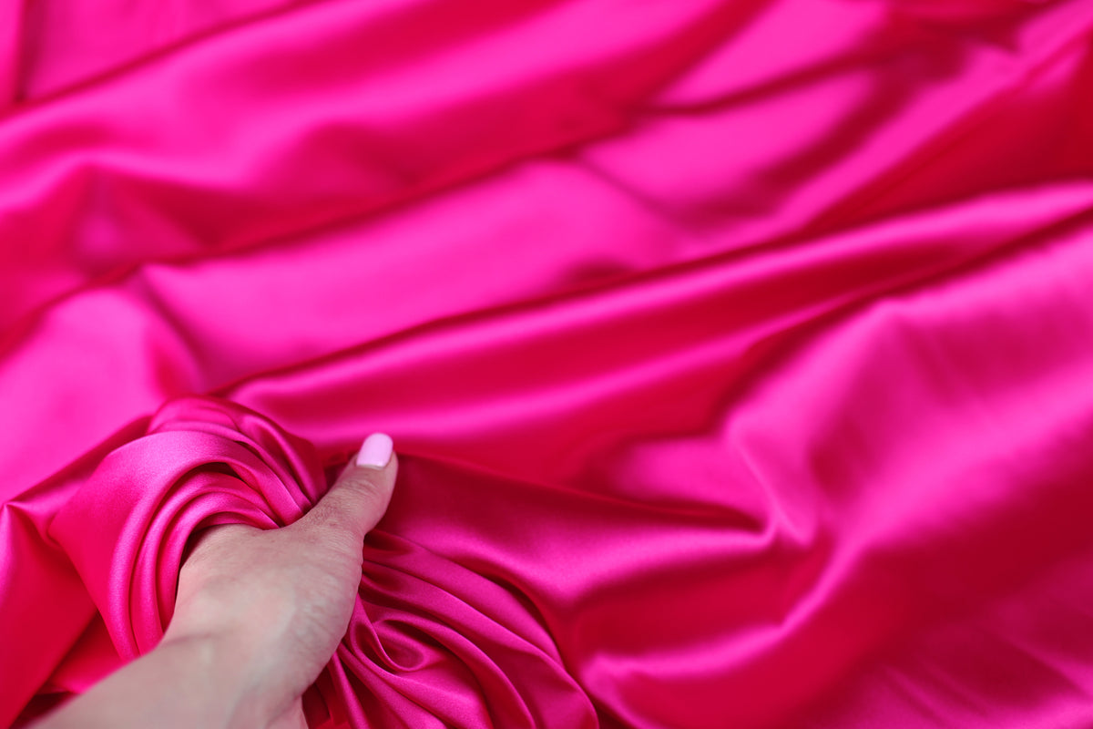 A hand is holding a piece of bright pink satin fabric, which appears to be soft and smooth.