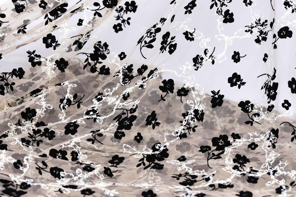 a close-up view of a fabric with a black and white floral pattern. The fabric appears to be made of sheer material, with the black flowers and leaves contrasting against the white background. The pattern is intricate and detailed, with the flowers and leaves filling the entire visible area of the fabric.