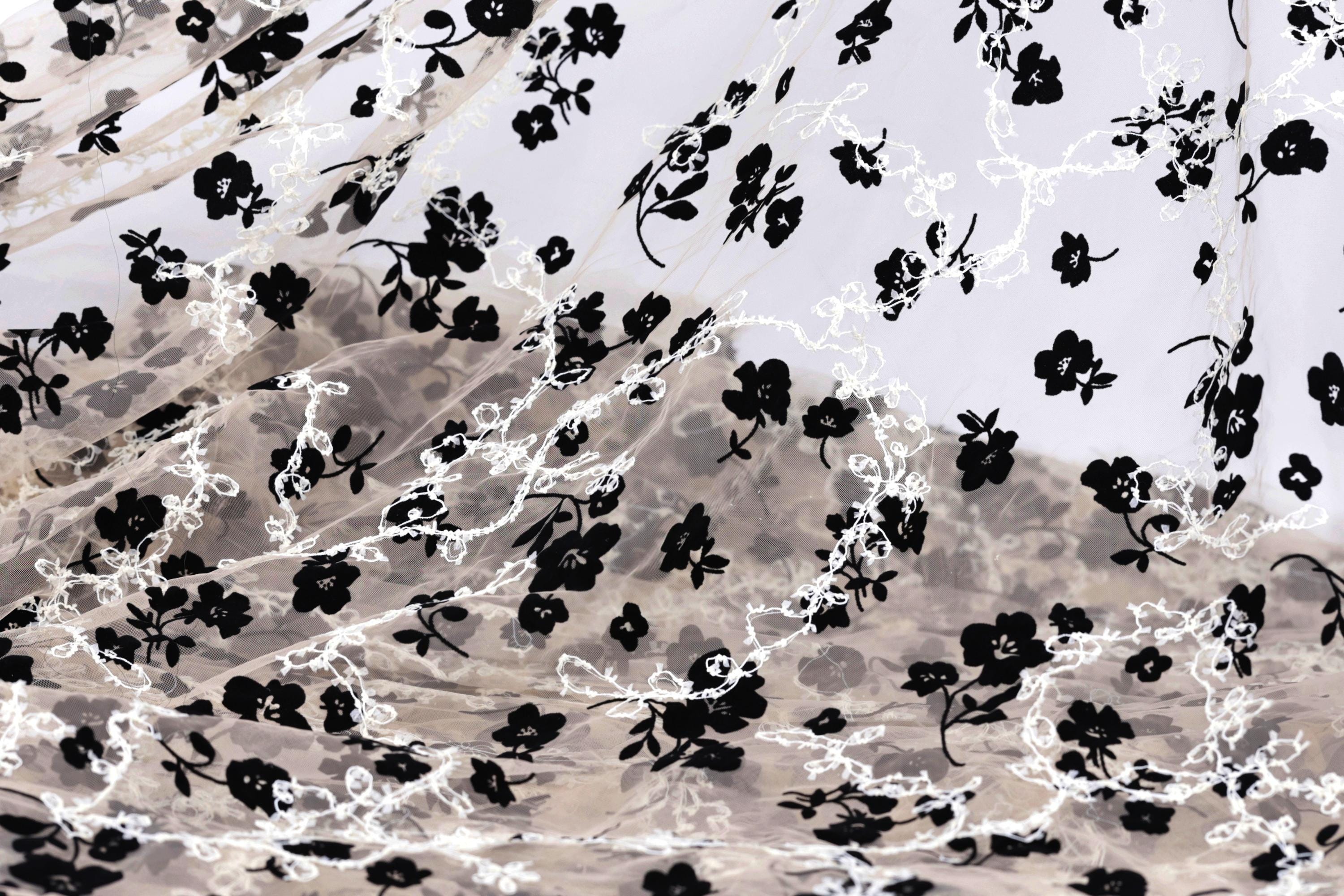 a close-up view of a fabric with a black and white floral pattern. The fabric appears to be made of sheer material, with the black flowers and leaves contrasting against the white background. The pattern is intricate and detailed, with the flowers and leaves filling the entire visible area of the fabric.
