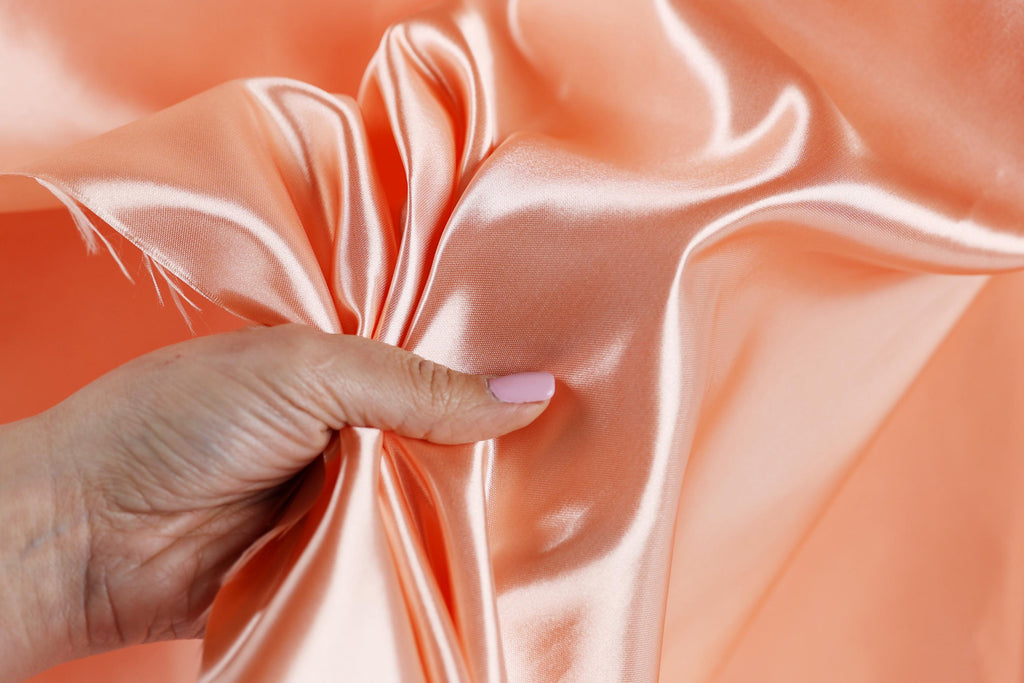 A hand is holding a piece of peach-colored satin fabric, with the fabric partially draped over the hand.