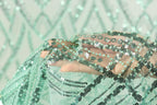 A hand is holding a piece of green and black sequin fabric, with the fabric's intricate patterns and designs visible.