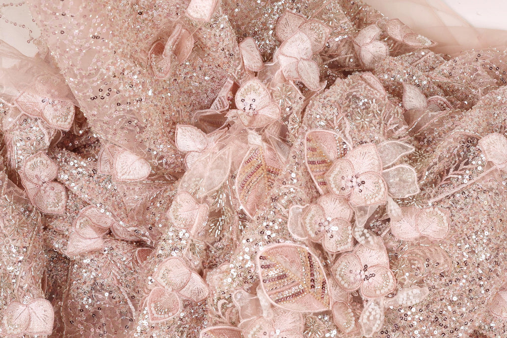 The image is a close-up of a delicate, intricately designed fabric with a floral pattern. The fabric is adorned with numerous small, pink flowers and sequins, creating a visually appealing and detailed appearance.